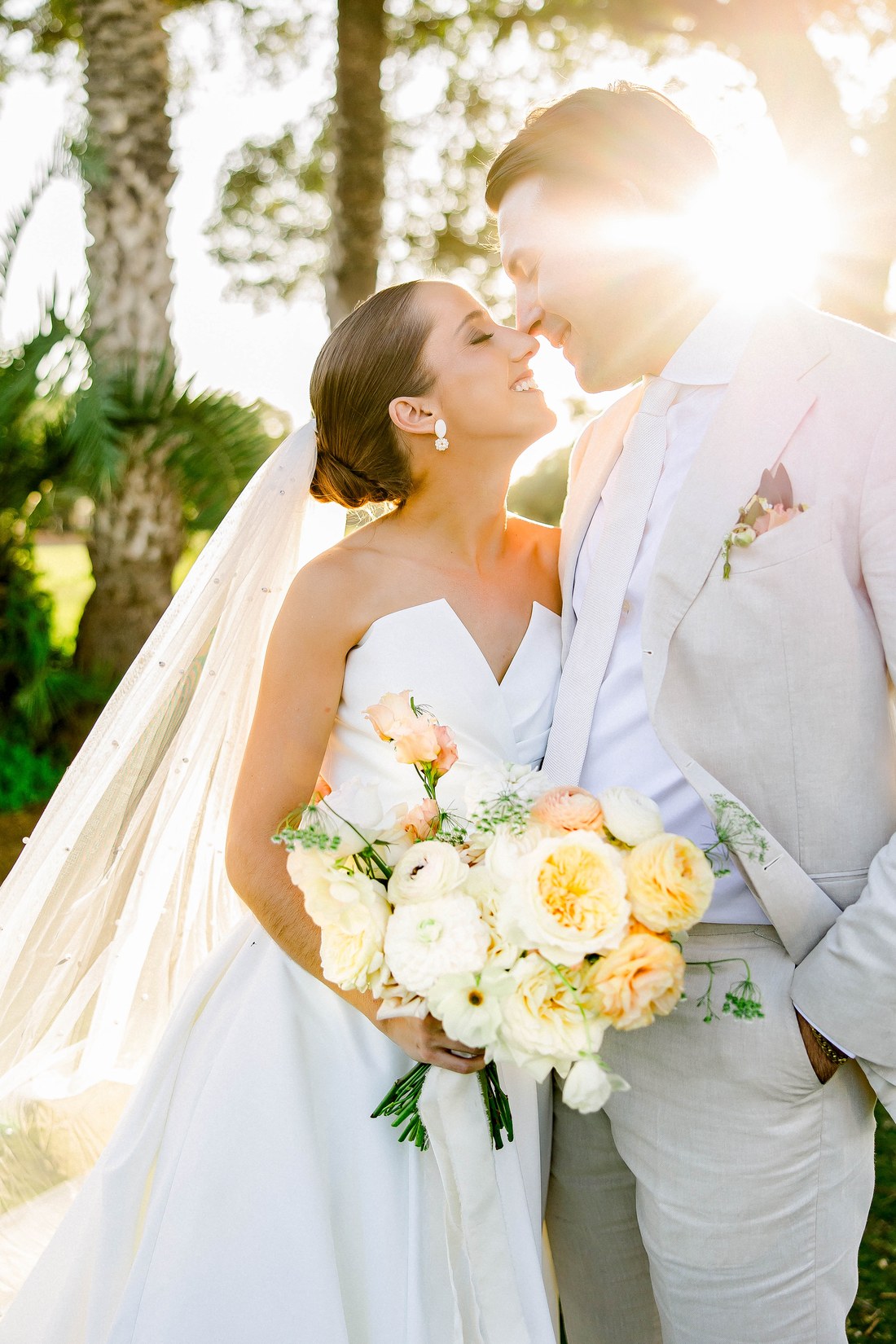 Couple sharing a golden hour moment among sun-drenched palms, bride in a flowing white gown with a soft peach bouquet — Tampa Bay wedding photographer Ailyn La Torre