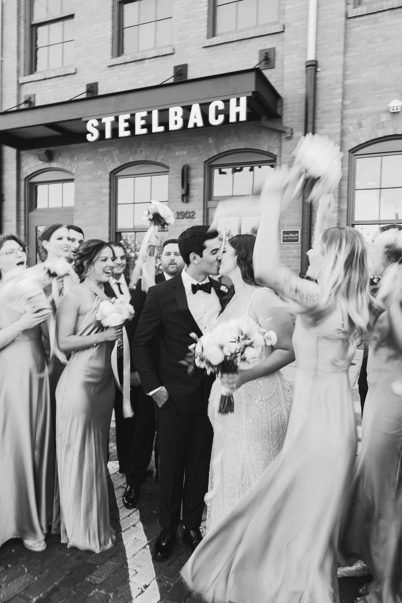 Bride and groom sharing a celebratory kiss outside Steelbach at Armature Works in Tampa, surrounded by a cheering bridal party — Tampa Bay wedding photographer Ailyn La Torre