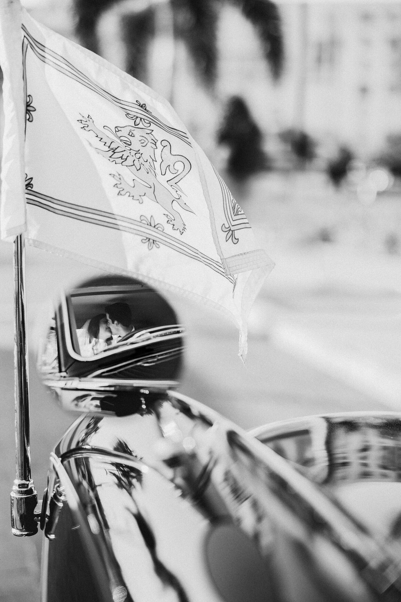 Couple reflected in the chrome mirror of a vintage car beneath a decorative heraldic flag — Tampa Bay wedding photographer Ailyn La Torre