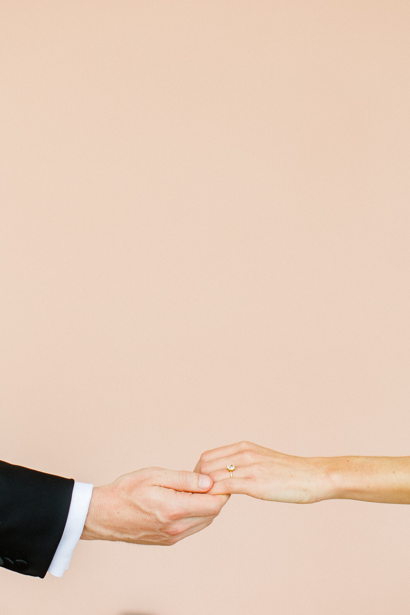 Groom reaching for his bride's hand, her gold solitaire engagement ring catching the light against a soft blush background — Tampa Bay wedding photographer Ailyn La Torre