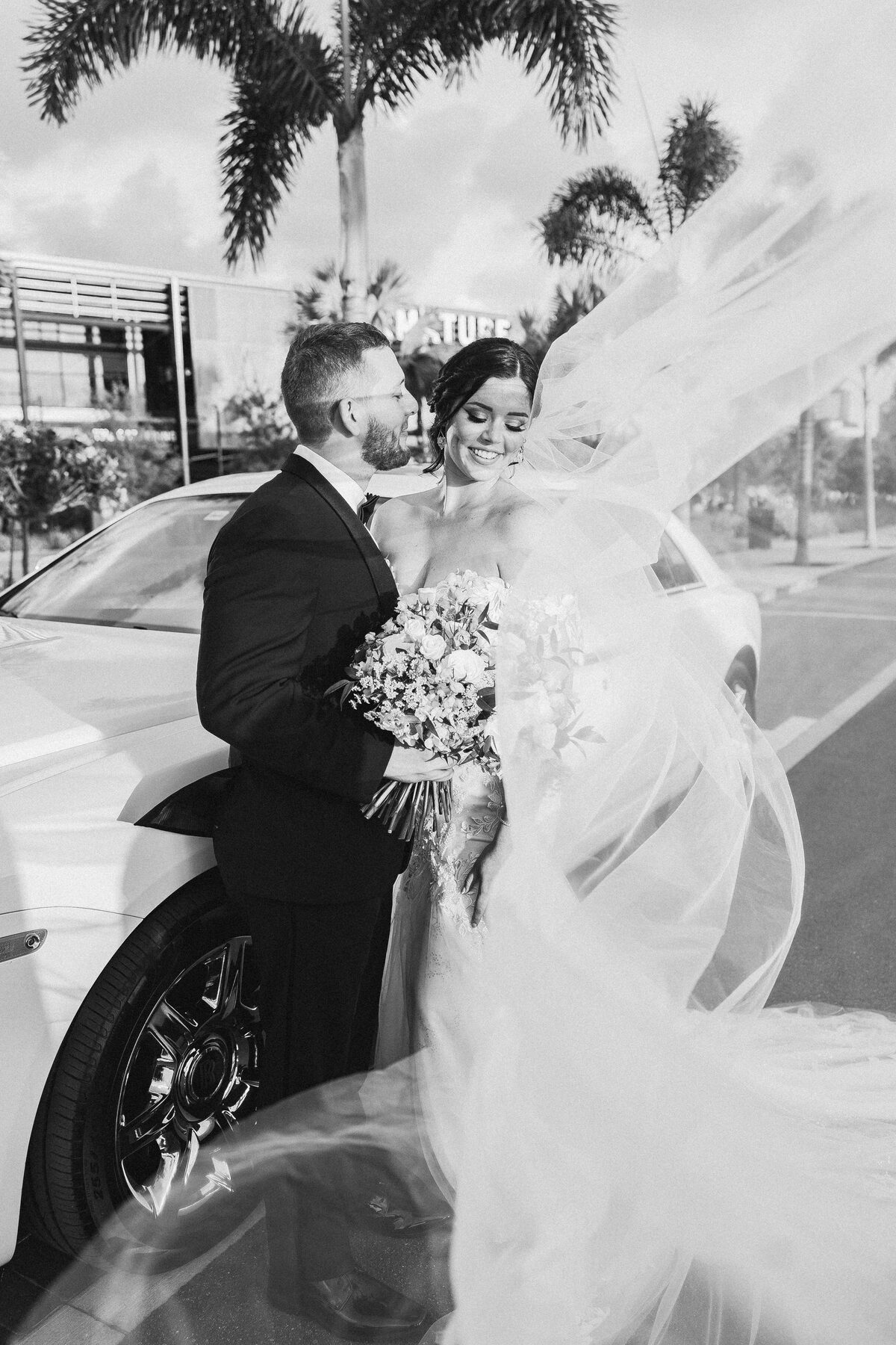 Bride's veil sweeping dramatically around a couple's portrait outside Armature Works in Tampa, groom kissing her cheek — luxury wedding photographer Ailyn La Torre