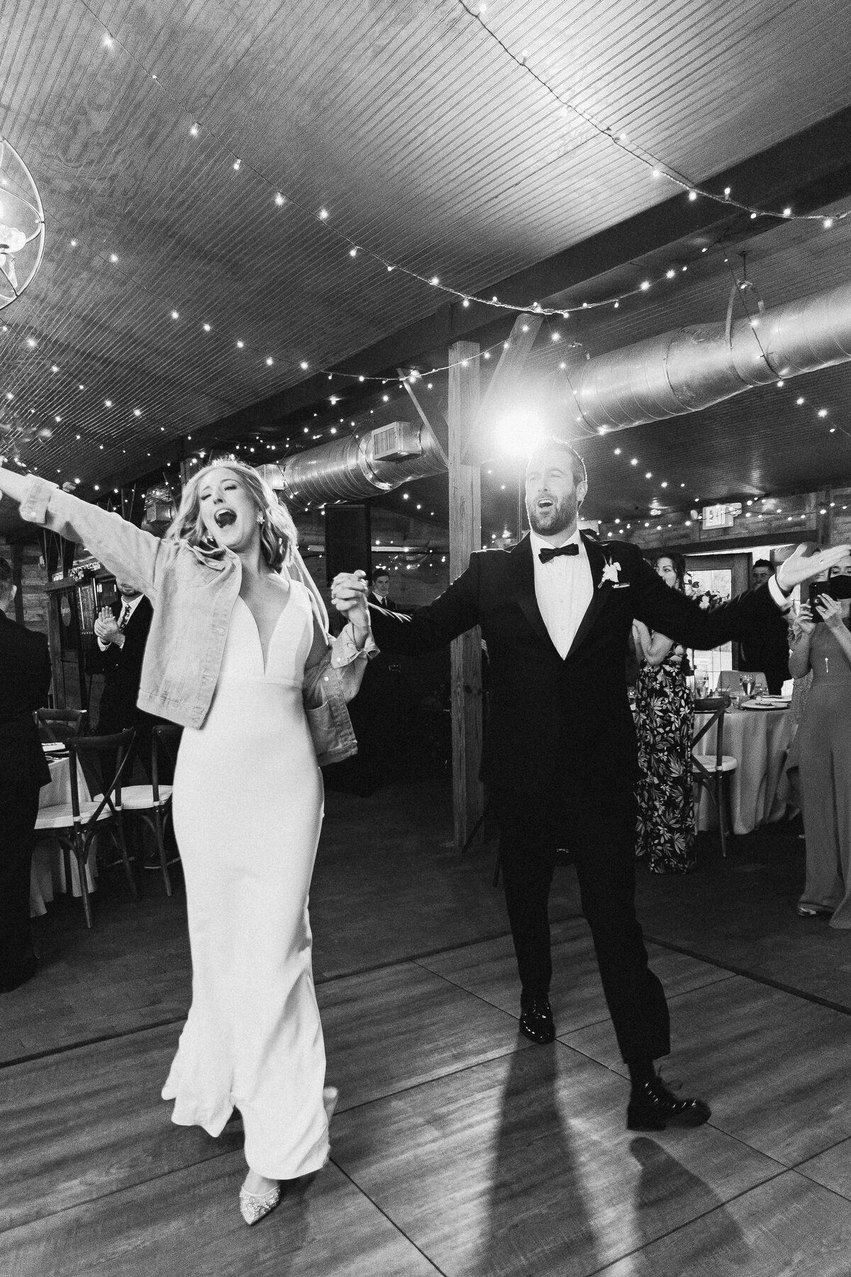 Bride in a sleek gown and denim jacket making a grand entrance with her groom at Cross Creek Ranch, arms outstretched under string lights — luxury wedding photographer Ailyn La Torre
