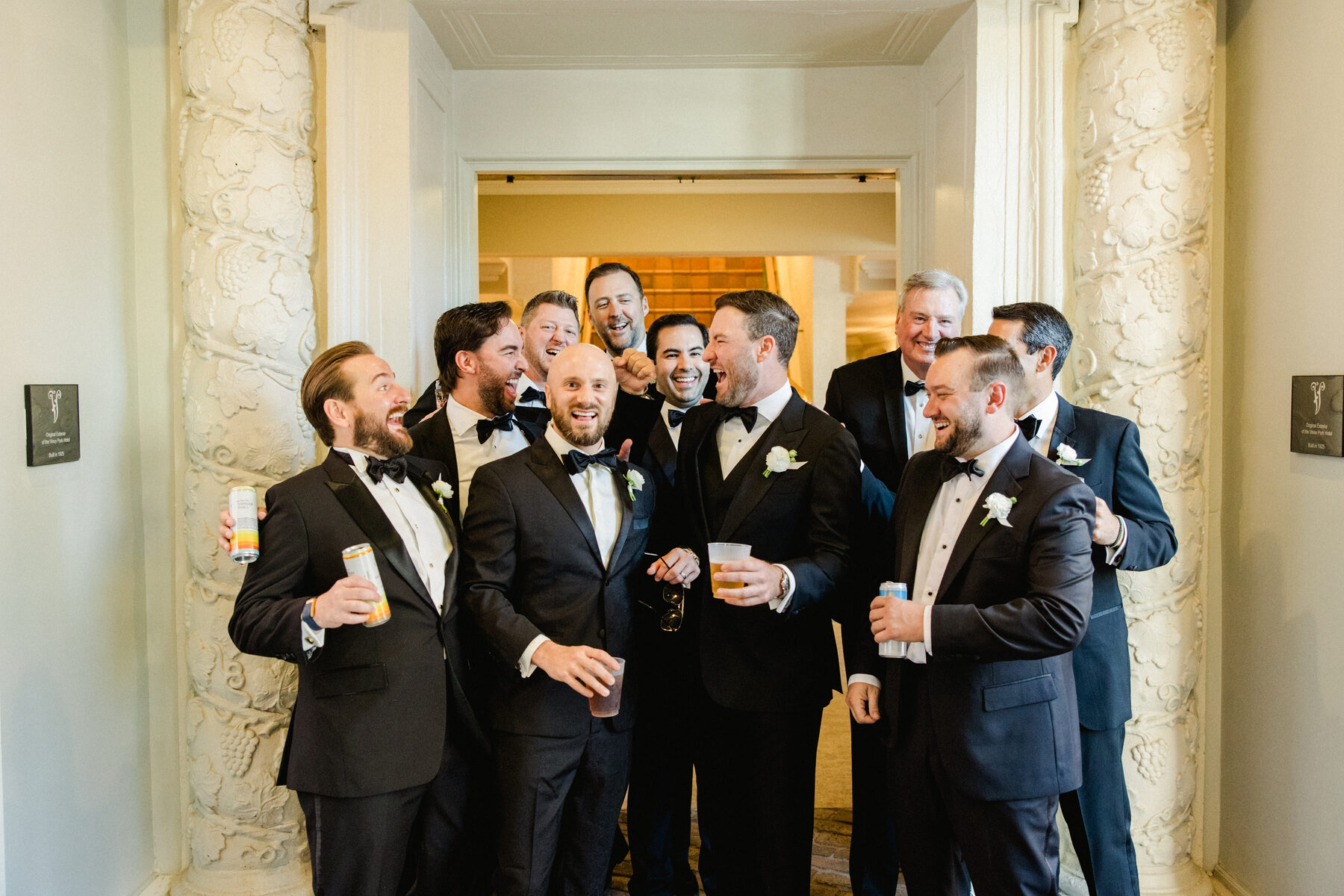 Groom and groomsmen laughing together in an ornate white hallway, drinks in hand, before a destination wedding ceremony — luxury wedding photographer Ailyn La Torre