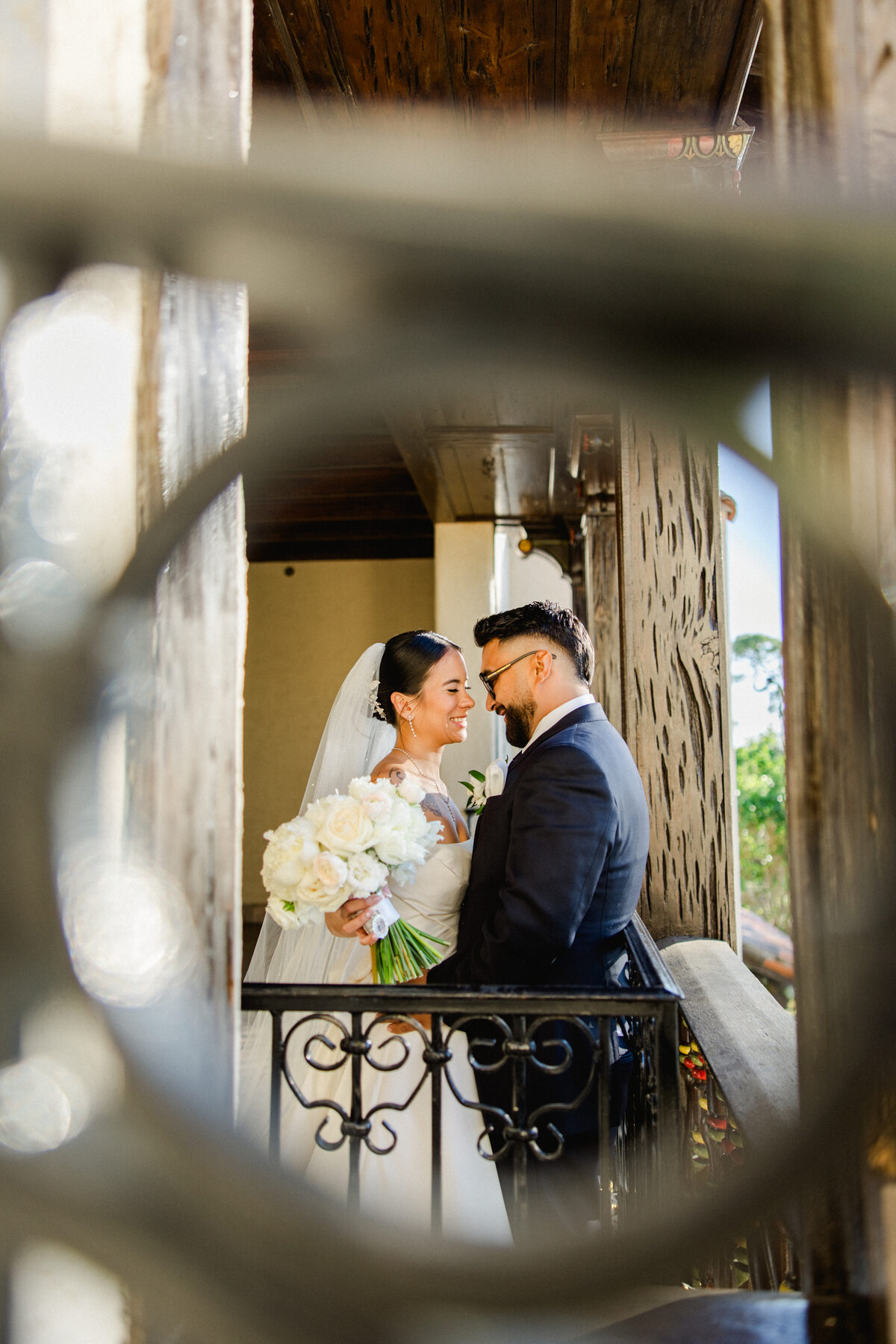 Bride and groom sharing a tender moment on the ornate balcony at Powel Crosley Estate, framed through decorative ironwork — luxury wedding photographer Ailyn La Torre